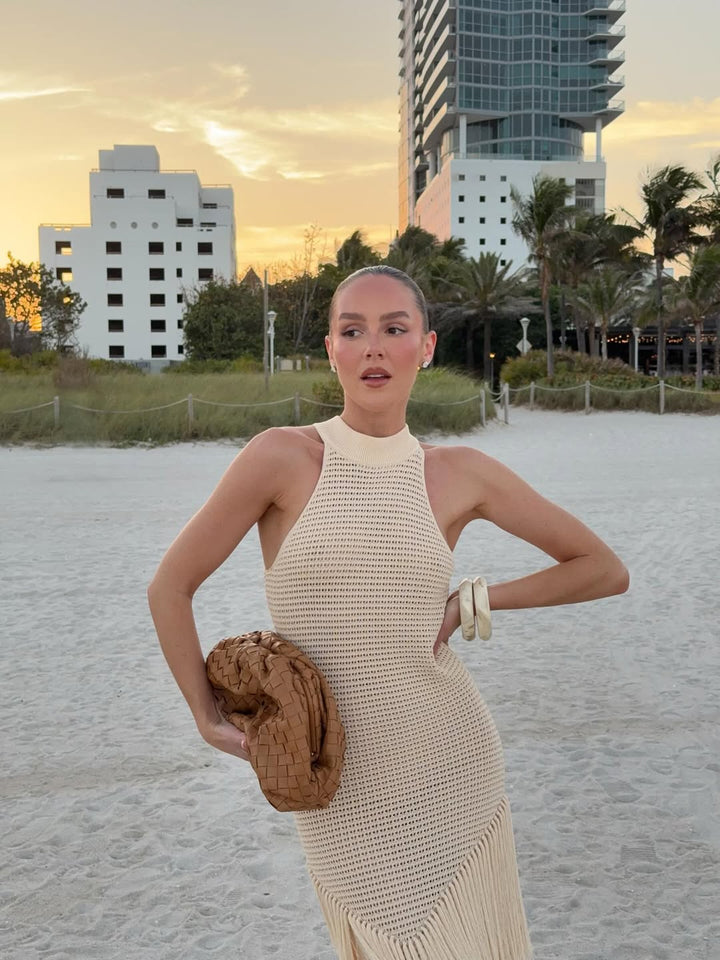 Woman in a beige dress standing on a beach with buildings and palm trees in the background
