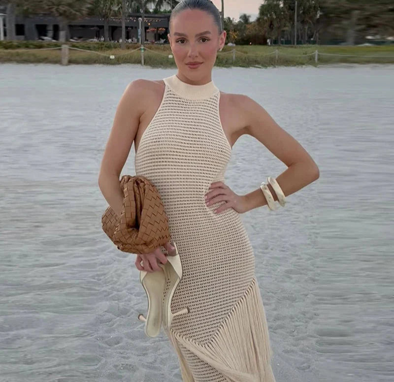 Woman in a beige dress standing on a beach holding a woven bag.